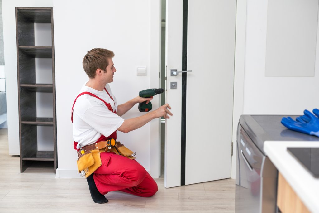Repair Service Concept. Portrait of smiling handsome young locksmith in blue uniform installing door knob on front wooden entrance door at home or office workplace.